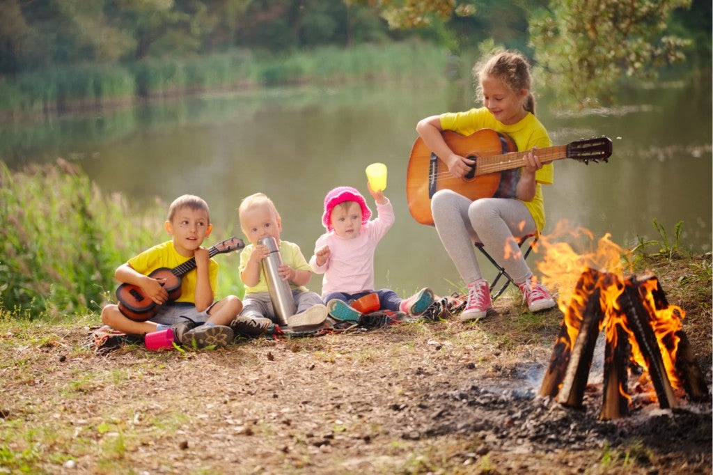 four children sit by a lake and a fire singing camp songs